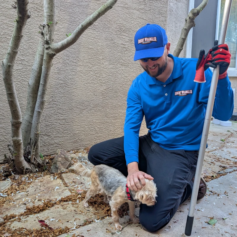 Man in a blue uniform kneels outside, gently stroking a small dog by a tree and a rake.